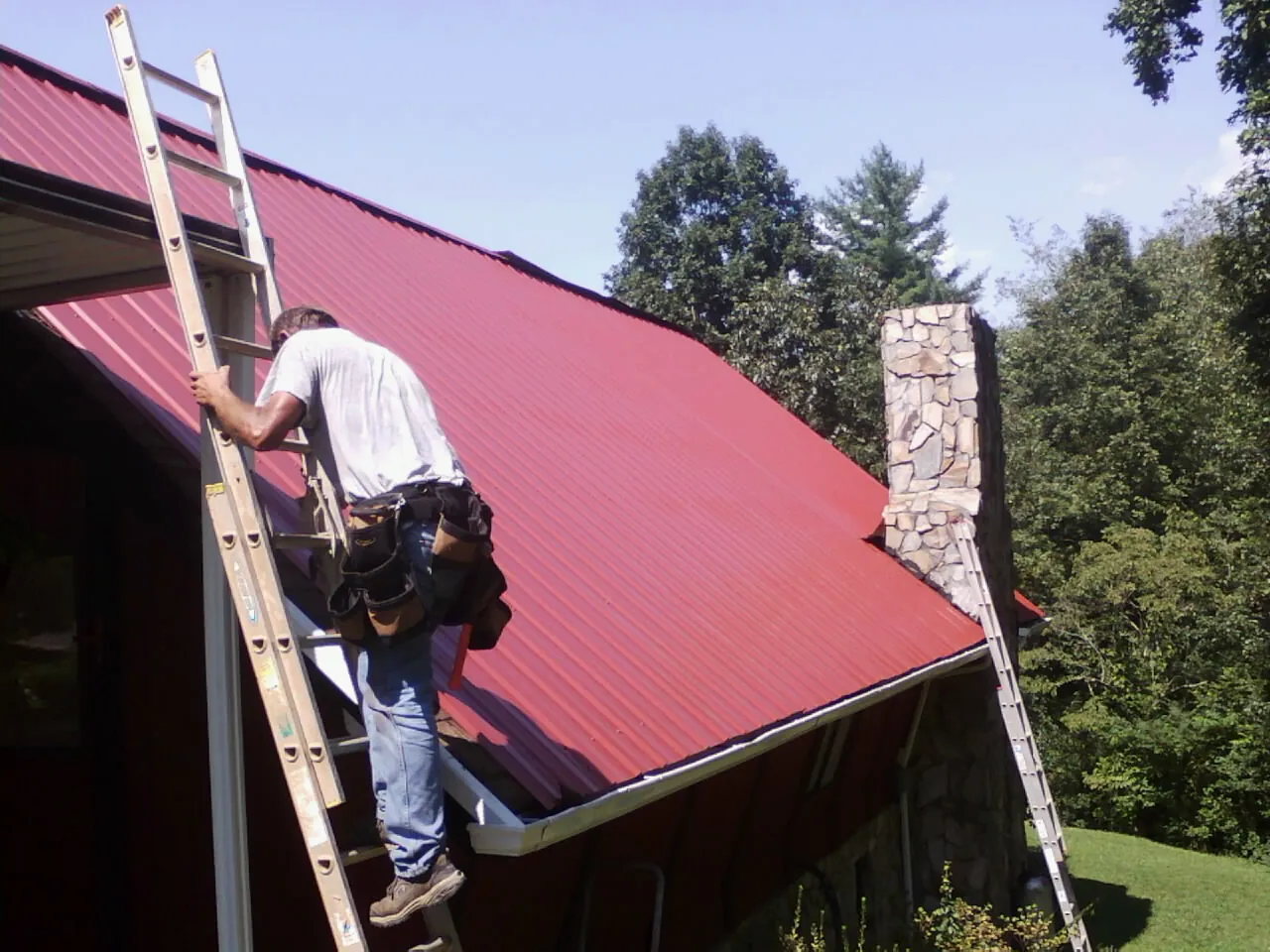 Roofer on a ladder beside a red metal roof and stone chimney — Soffit & Fascia Repair by Roofing Brothers in Manitou Springs